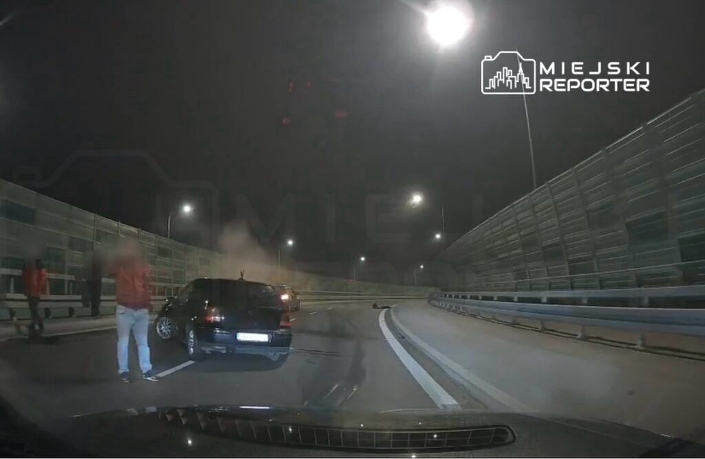 Grupa osób w czerwonych kurtkach stoi obok uszkodzonego samochodu na drodze, w tle widać dym.