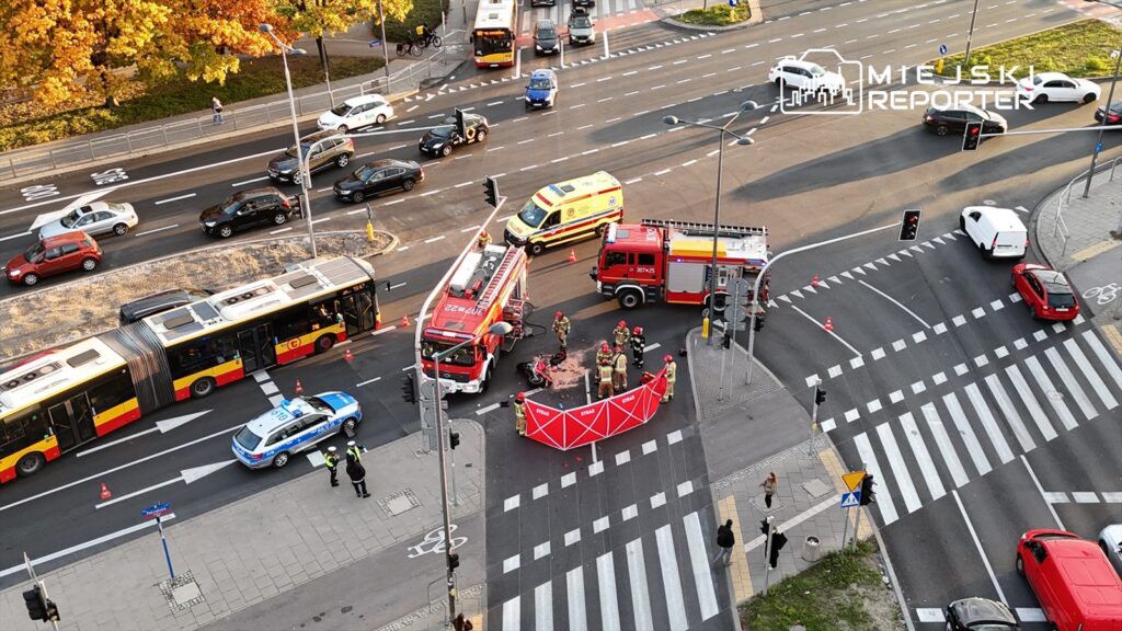 Grupa strażaków oraz ratowników medycznych pracuje przy wypadku drogowym, otoczona czerwonym parawanem, na ruchliwej ulicy.