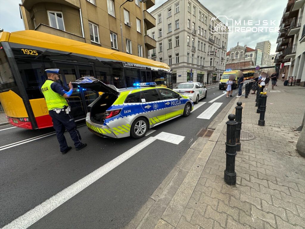 Policjant w kamizelce odblaskowej przeszukuje bagażnik radiowozu na ruchliwej ulicy, obok zatrzymany autobus.