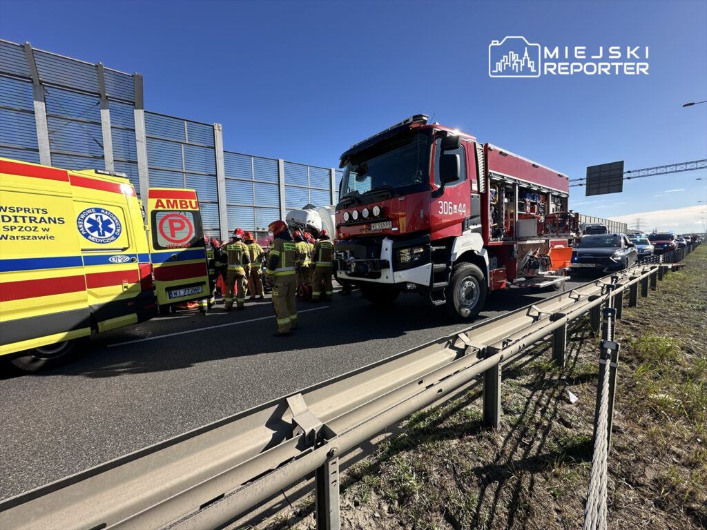 Zespół ratunkowy w mundurach pracuje przy wypadku drogowym na autostradzie, obok wozu strażackiego i karetki.