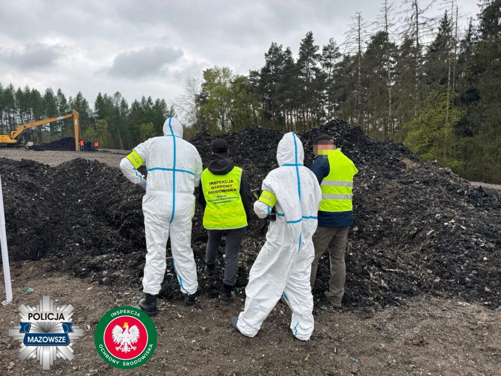 Grupa pracowników w kombinezonach ochronnych obserwuje prace przy rozładunku odpadów w lesie.