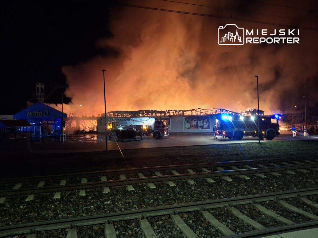 Pożar strawił dach supermarketu Netto, a w tle widać płonące fragmenty budynku oraz wozy strażackie.