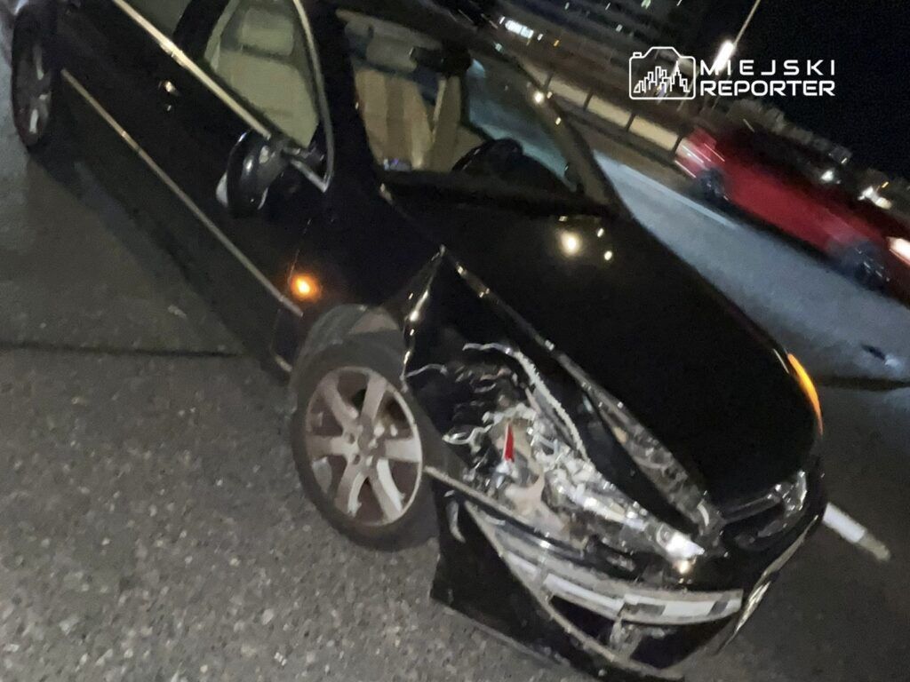 Czarny samochód osobowy z uszkodzoną przednią częścią stoi na drodze w nocy. W tle widać inne pojazdy.