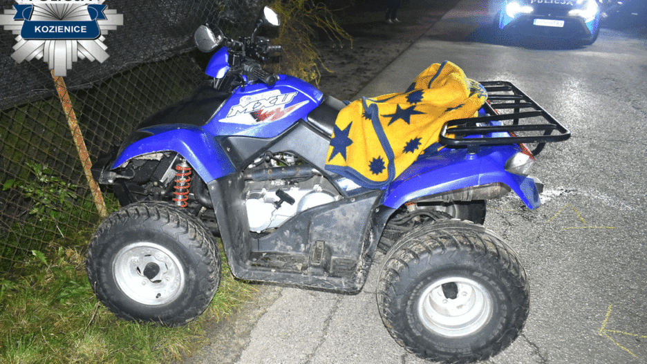 Niebieski quad zaparkowany na poboczu drogi, przykryty żółtym kocem w gwiazdy, obok policyjny radiowóz.