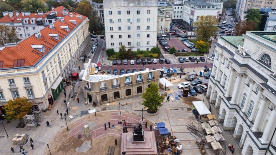 Widok na plac z pomnikiem, w tle nowoczesne budynki i zieleń, w centrum trwają prace budowlane.