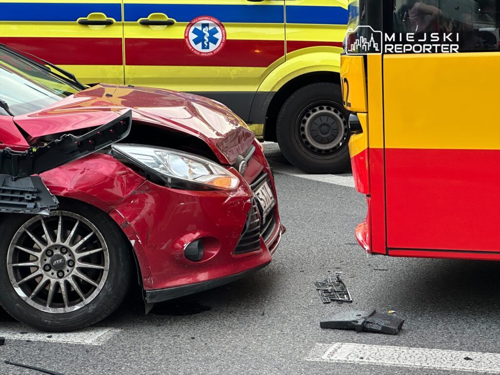 Czerwony samochód osobowy z uszkodzoną maską zderza się z żółtym autobusem, obok stoją karetki pogotowia.