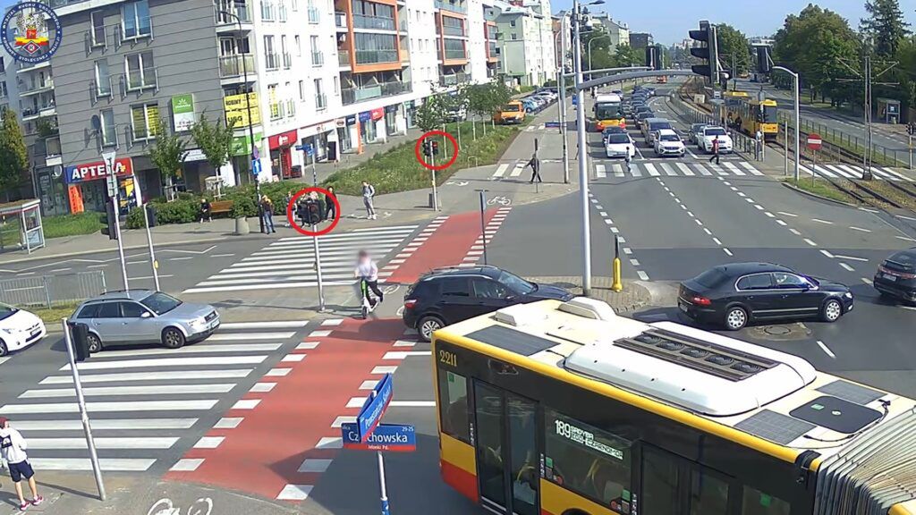 Grupa osób stoi na przejściu dla pieszych, a w tle ruchliwa ulica z samochodami i autobusem.
