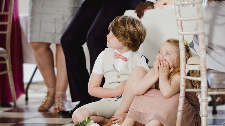 Children are sitting on the dancefloor by a table at a wedding. They are watching the bride and groom share their first dance. DEAN HINDMARCHchildren, little boy, little girl, sitting, siblings, brother, sister, watching, spectating, wedding guest, pageboy, flower girl, bridesmaid, 5 years, 9 years, caucasian, relaxation, peaceful, enjoyment, stressfree, caucasian, british culture, wedding, wedding reception, wedding ceremony, marriage, celebration, celebratory event, tiled floor, dancefloor, formal, well dressed, childhood, youth culture, cute, family, friends, fun, indoors, cute, innocent, purity, young, lifestyles, weekend activity, daughter, son, brother, sister, tranquil scene