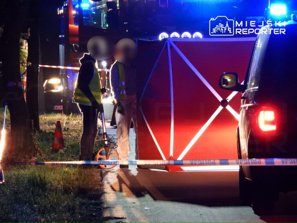 Na zdjęciu widoczna jest scena wypadku drogowego, gdzie służby ratunkowe prowadzą działania. Policjanci zabezpieczają teren z użyciem taśmy policyjnej, a strażacy i ratownicy pracują nad ustaleniem okoliczności zdarzenia. W tle widać pojazdy służbowe i oświetlenie.