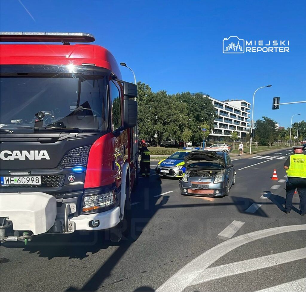 Czerwony wóz strażacki stoi obok uszkodzonego samochodu na skrzyżowaniu, obok policyjny radiowóz.