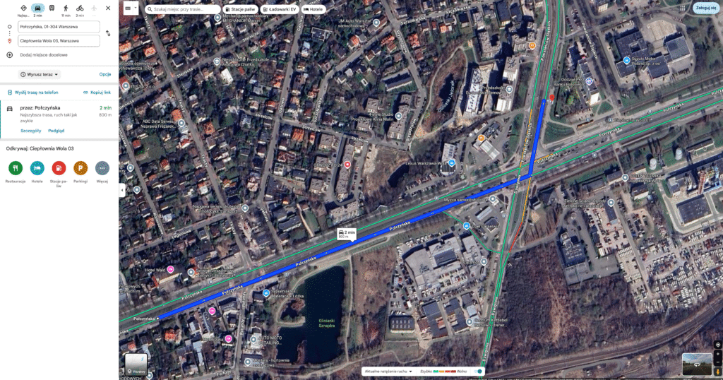 Widok satelitarny obszaru miejskiego z drogami, budynkami i parkingami, w tym zbiornikiem wodnym w dolnej części obrazu.