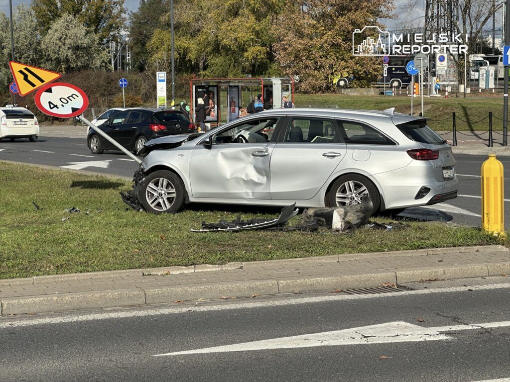 Uszkodzony srebrny samochód osobowy zaparkowany na trawie obok znaku drogowym informującym o ograniczeniu wysokości do 4 m.