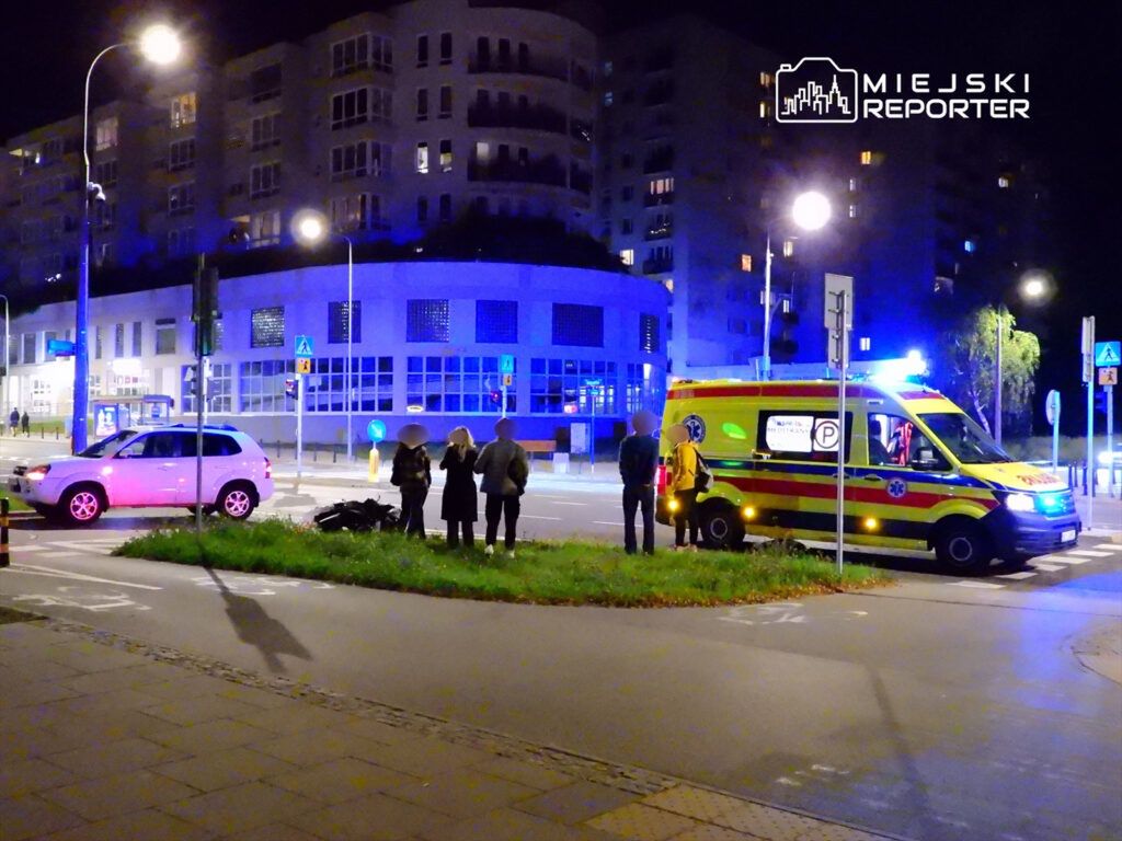 Grupa osób stoi obok ambulansu na skrzyżowaniu, a w tle widać oświetlone budynki nocą.
