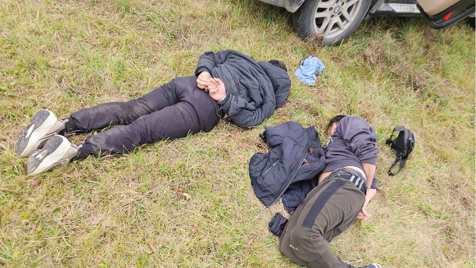 Dwie osoby leżą na trawie obok samochodu, jedna z nich ma ręce związane, obie w ciemnych ubraniach.