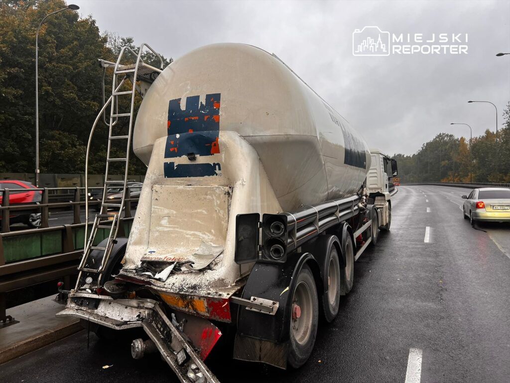 Ciągnik siodłowy z cysterną stoi na drodze, obok niego przejeżdża samochód osobowy w deszczowej aurze.