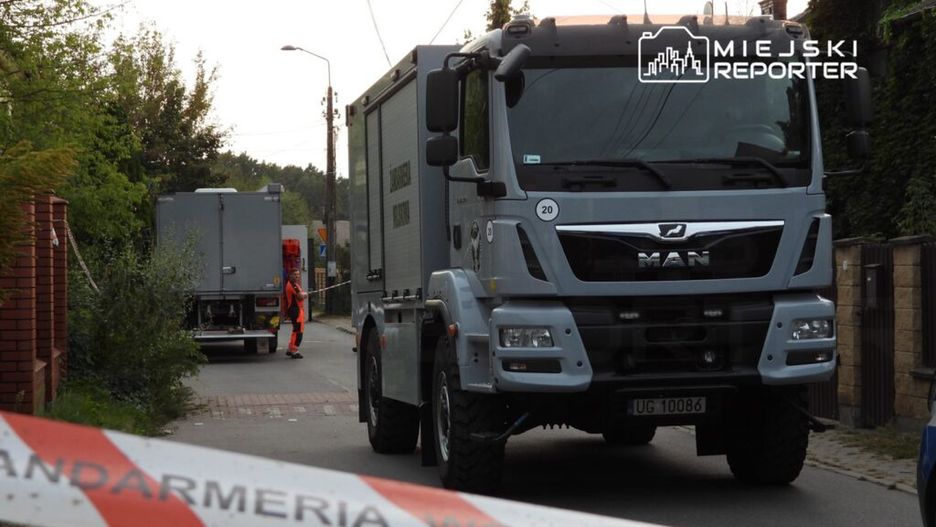 Na ulicy stoi ciężarówka MAN oraz pojazd z kontenerem, a pracownik w pomarańczowym uniformie zabezpiecza teren taśmą.