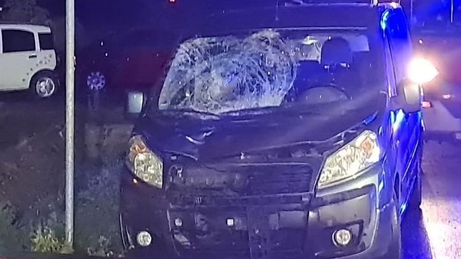 Rozbity czarny van z uszkodzoną przednią szybą stoi na poboczu drogi, obok znaku drogowym. W tle widać niebieskie światła pojazdów służb ratunkowych.