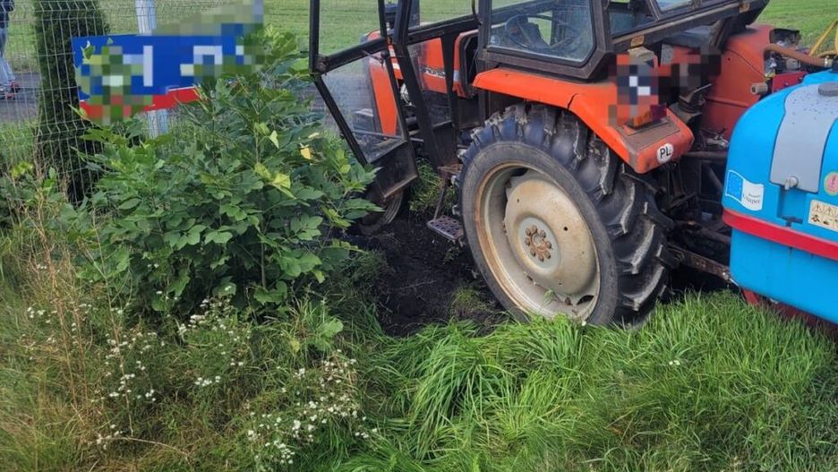 Traktor z czerwoną kabiną stoi w zielonej okolicy, obok ogrodzenia i krzewów.