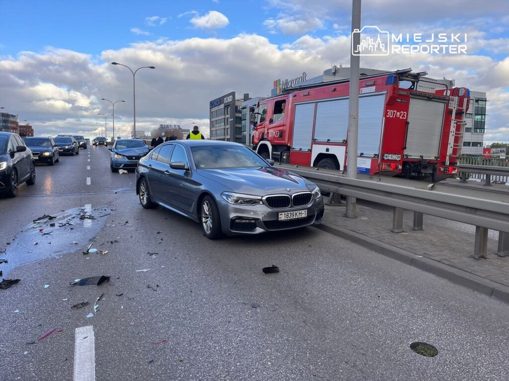 Kolizja drogowa z udziałem szarego BMW oraz uszkodzonymi pojazdami na ruchliwej ulicy, w tle wóz strażacki.