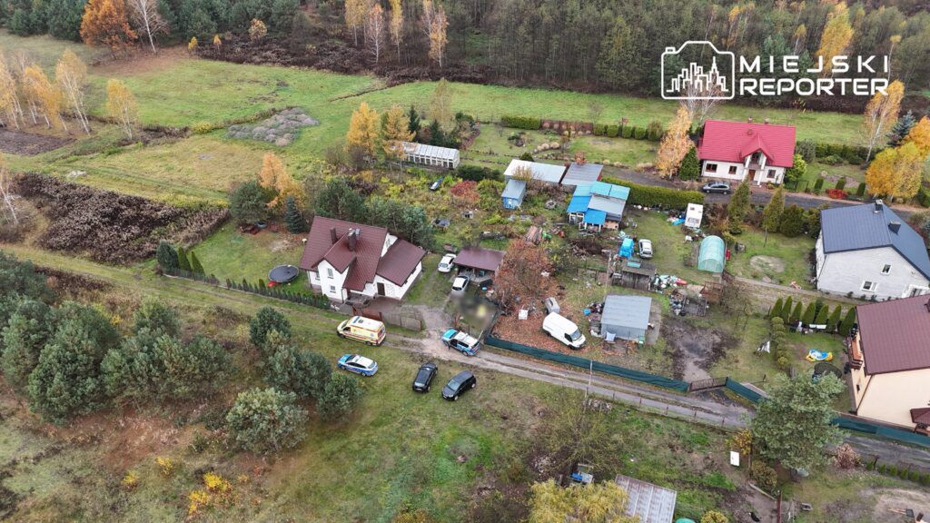 Widok z góry na osiedle domków jednorodzinnych z samochodami, w tym karetka pogotowia i radiowóz, w otoczeniu zieleni.