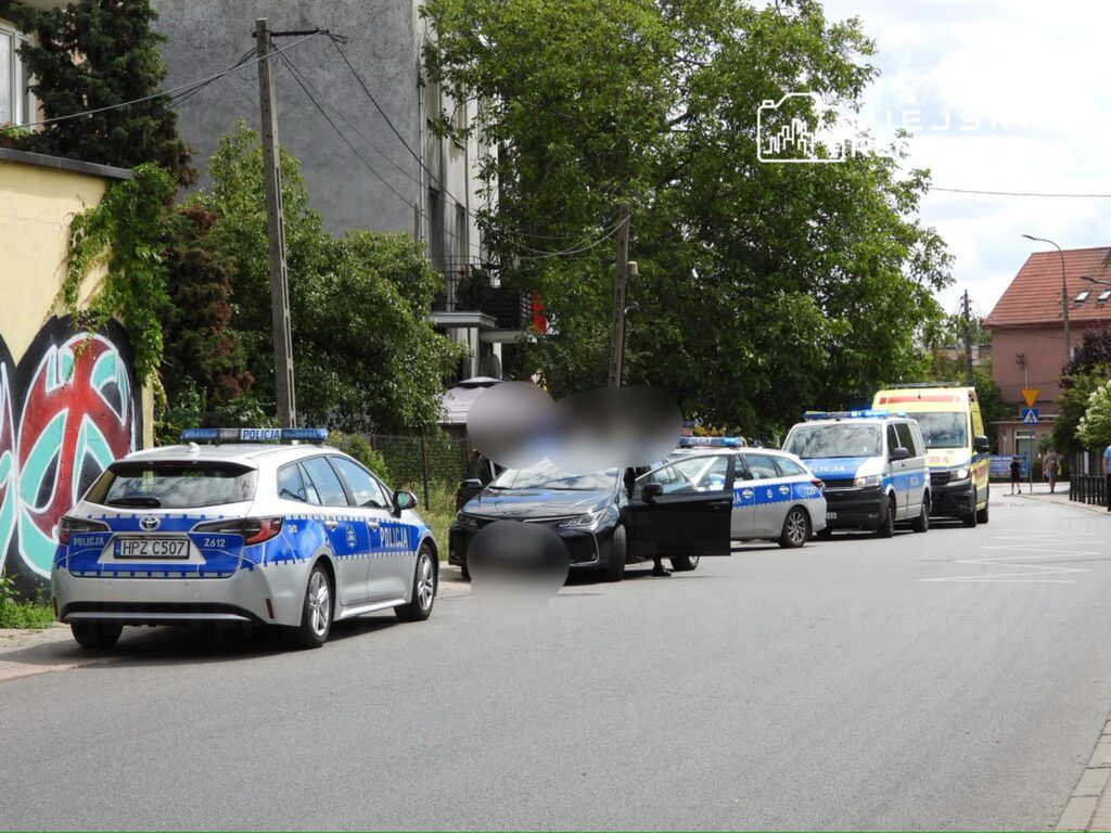 Grupa radiowozów policyjnych i karetka stojące na ulicy, obok graffiti na ścianie budynku.