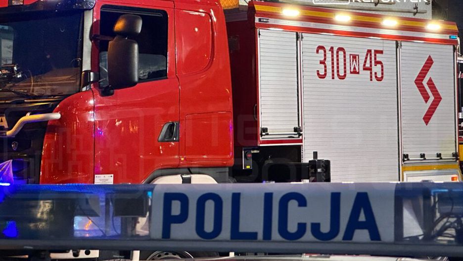 Czerwony wóz strażacki i radiowóz policyjny zaparkowane na nocnej ulicy, oświetlone niebieskimi światłami.