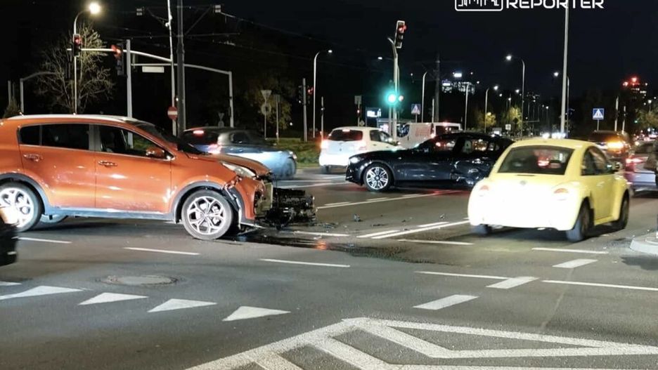 Zderzenie dwóch samochodów na skrzyżowaniu, z uszkodzonym pomarańczowym pojazdem i żółtym autem przejeżdżającym obok.
