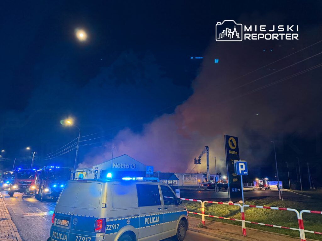 Strażacy gaszą pożar w pobliżu budynku sklepu Netto, w otoczeniu wozów policyjnych i strażackich w nocy.