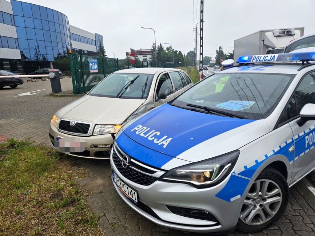 Policyjny radiowóz zaparkowany obok uszkodzonego samochodu na parkingu przed nowoczesnym biurowcem.