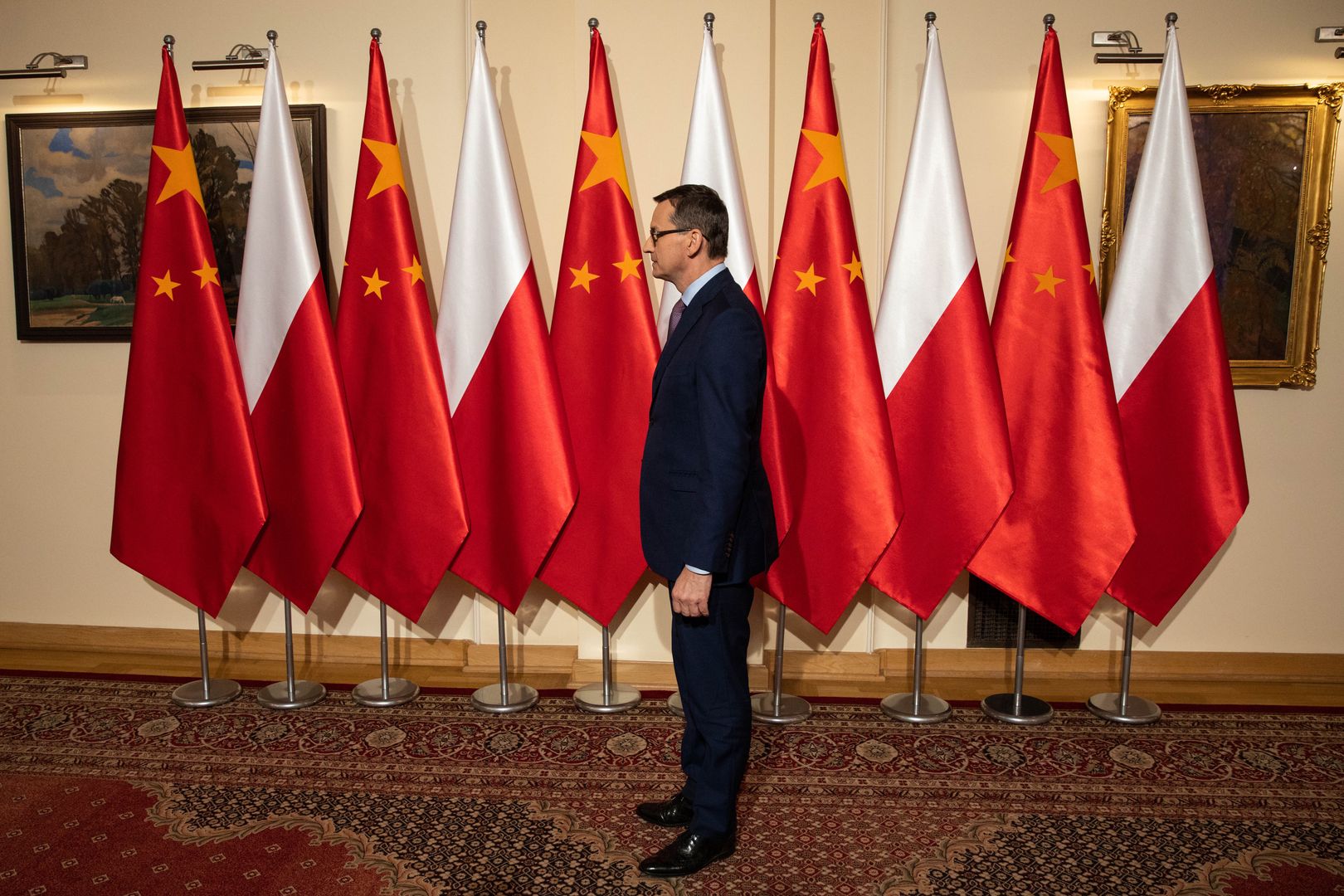 Premier Mateusz Morawiecki na tle flag polskiej i chińskiej.
