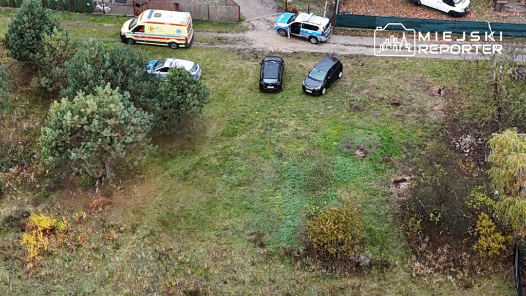Na trawie stoją trzy samochody: dwa czarne osobowe oraz nieoznakowany radiowóz, obok parkowana karetka pogotowia.