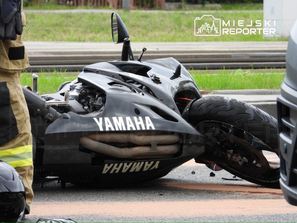Czarny motocykl marki Yamaha leży na boku na drodze, obok stoi strażak w mundurze.