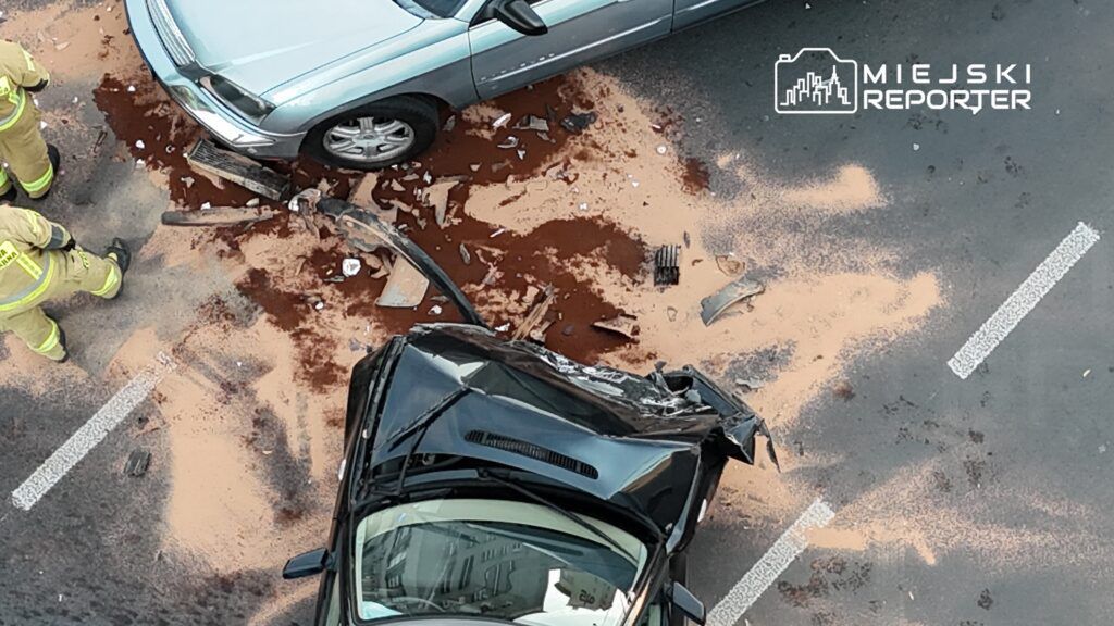 Czarny samochód z uszkodzoną karoserią leży na jezdni obok srebrnego pojazdu, wokół rozbite szkło i plamy oleju.