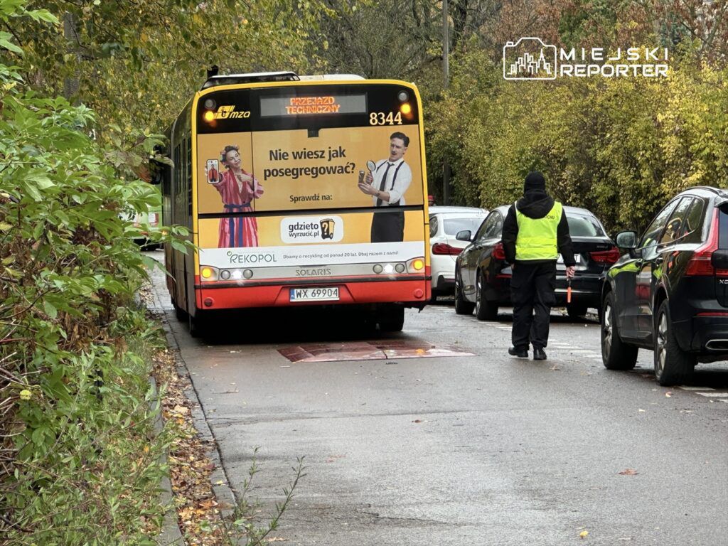 Autobus z reklamą na tylnej części zatrzymany na ulicy, obok pracownik w kamizelce odblaskowej kierujący ruchem.