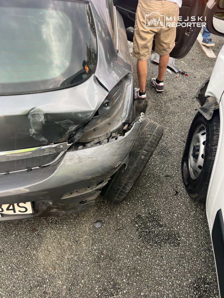 Uszkodzony samochód osobowy z wgniecioną tylną częścią stoi na parkingu, obok mężczyzna w krótkich spodniach bada uszkodzenia.