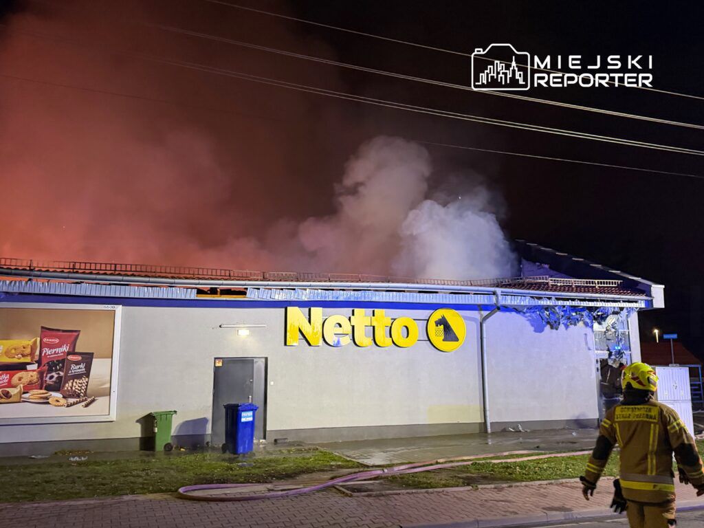 Pożar dachu supermarketu Netto, z którego wydobywa się dym, w otoczeniu ciemnej nocy.