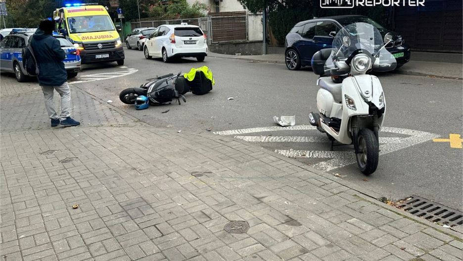 Mężczyzna w ciemnej kurtce obserwuje wypadek motocyklowy na ulicy, obok leży przewrócony skuter.