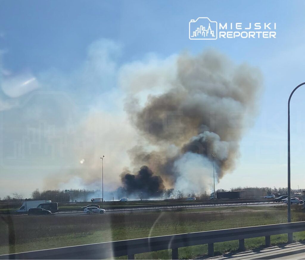 Gęsty dym unosi się nad drogą, obok której poruszają się samochody, w tle widać zalesiony teren.