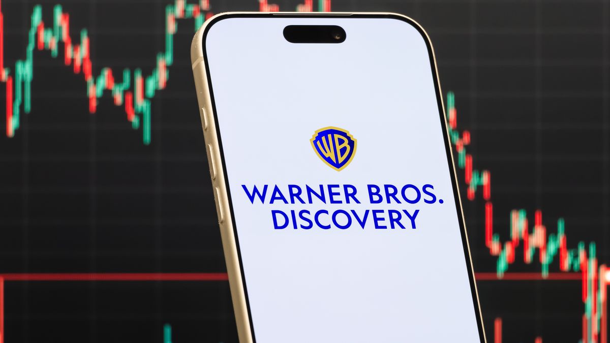 QIANJIANG, CHINA - FEBRUARY 15: In this photo illustration, a smartphone displays the logo of Warner Bros. Discovery, Inc. (NASDAQ: WBD), an American media and entertainment company operating film studios, television networks and streaming platforms, in front of a screen showing the company's latest stock market chart on February 15, 2026 in Qianjiang, Hubei Province, China. (Photo illustration by Cheng Xin/Getty Images)
