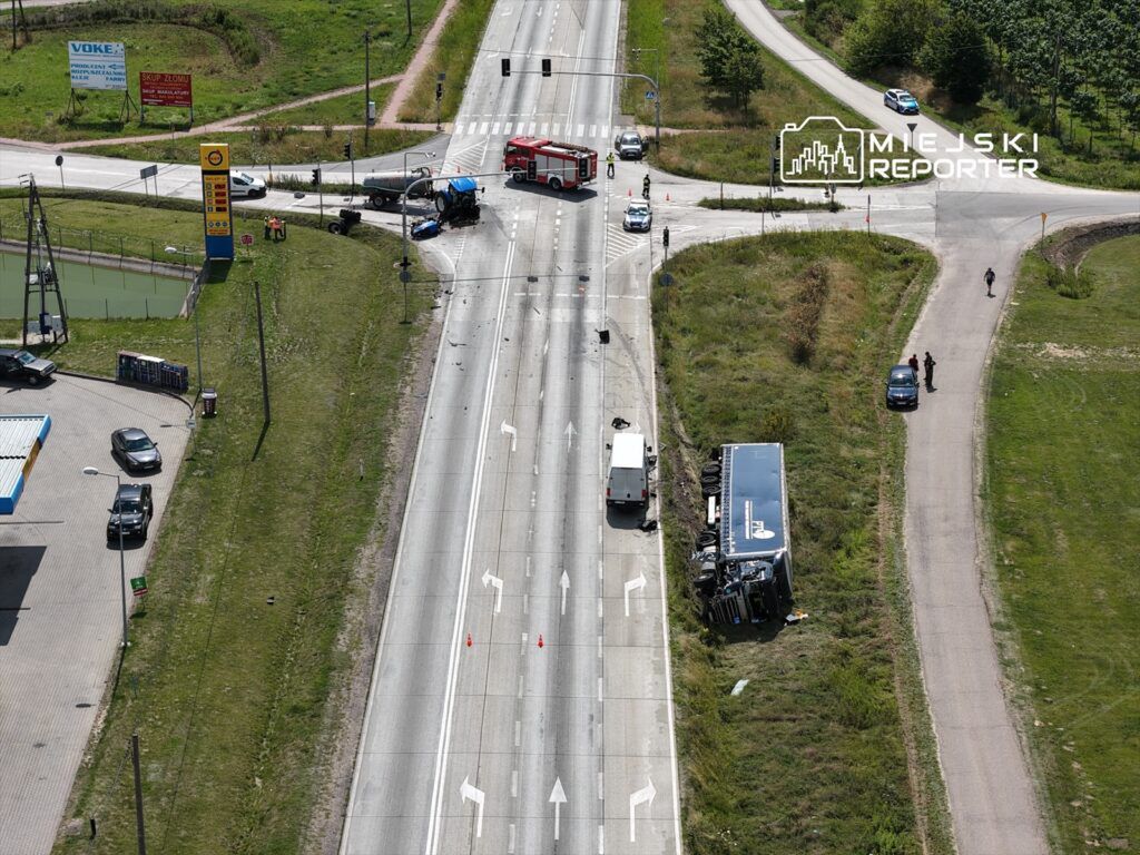 Wielka ciężarówka leży na boku obok drogi, obok niej kilka pojazdów i straż pożarna, na tle zielonego krajobrazu.