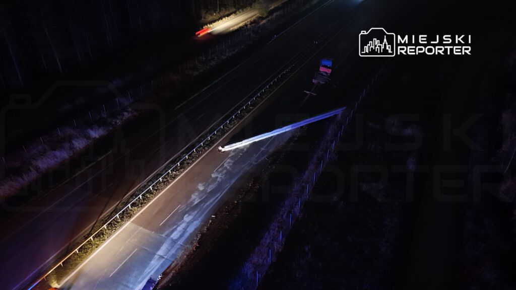 Na drodze krajowej widoczny wypadek, uszkodzony pojazd oraz rozbite barierki, otoczone przez ciemność lasu.