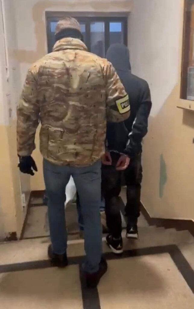 Funkcjonariusz w kamizelce policyjnej prowadzi zatrzymanego mężczyznę w ciemnym dresie przez korytarz.