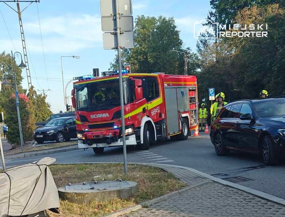 Czerwony wóz strażacki z włączonymi sygnałami świetlnymi stoi na skrzyżowaniu, obok grupy strażaków w odblaskowych kamizelkach.