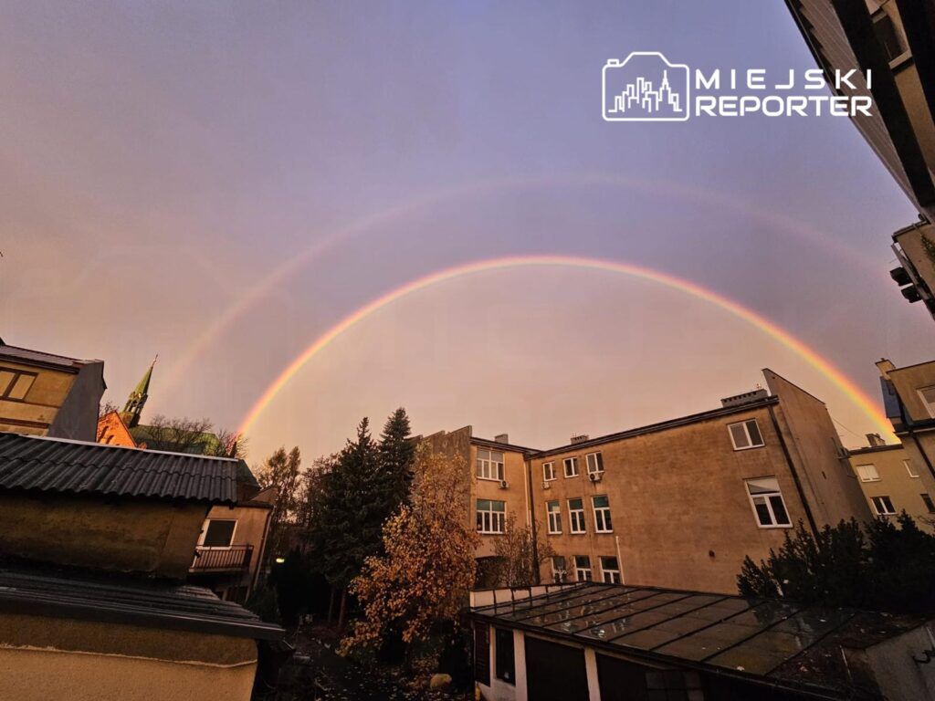 Podwójna tęcza widoczna na tle miejskich budynków i zielonych drzew w słoneczny dzień.