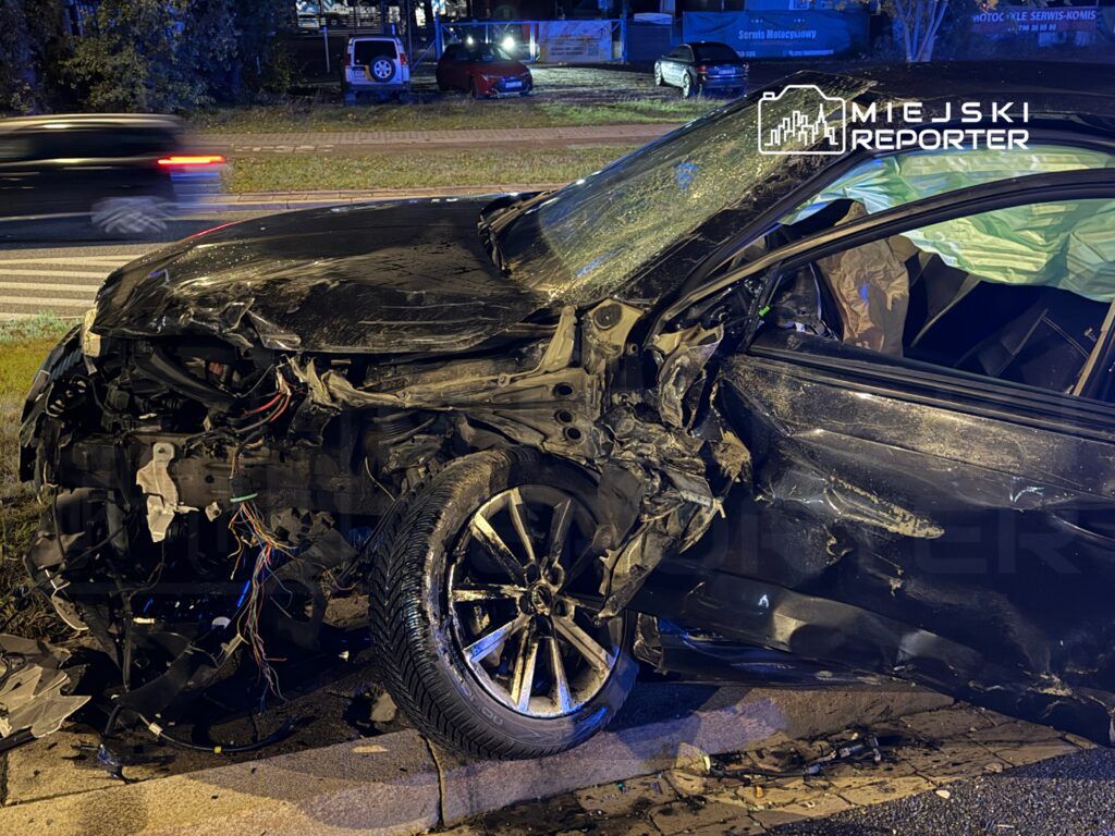 Zniszczony czarny samochód osobowy leży na poboczu drogi, z odkształconą karoserią i widocznymi przewodami elektrycznymi.