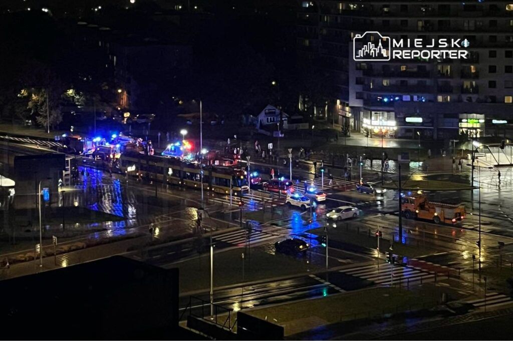 Na skrzyżowaniu ulic znajdują się pojazdy policyjne oraz straż pożarna z włączonymi sygnałami świetlnymi. Na mokrej nawierzchni widać odbicia świateł.