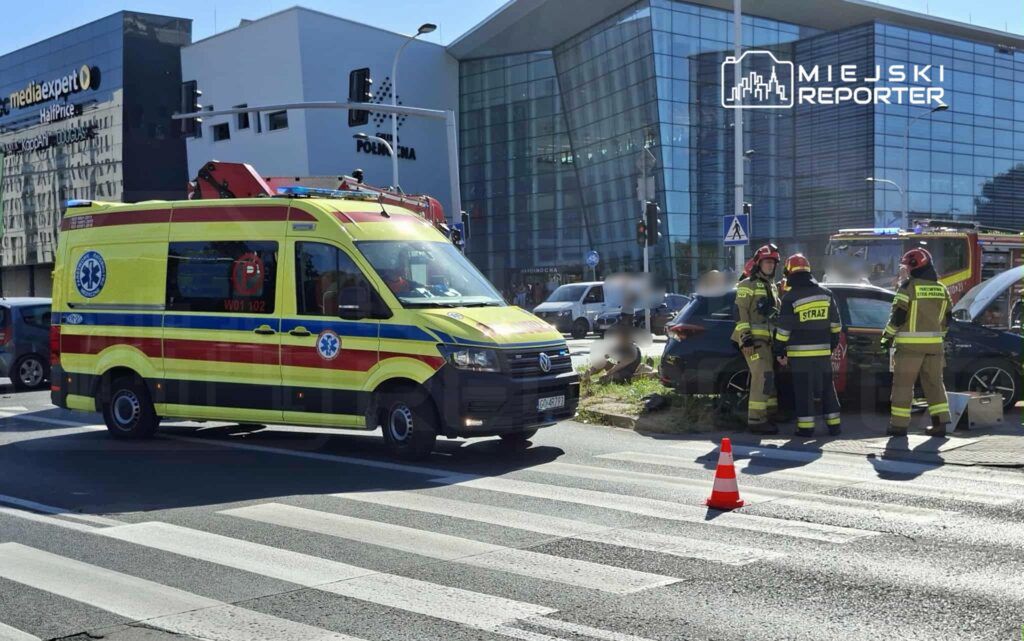 Zespół ratunkowy w żółto-czerwonym ambulansie zatrzymany przy wypadku drogowym; strażacy rozmawiają obok uszkodzonego samochodu.