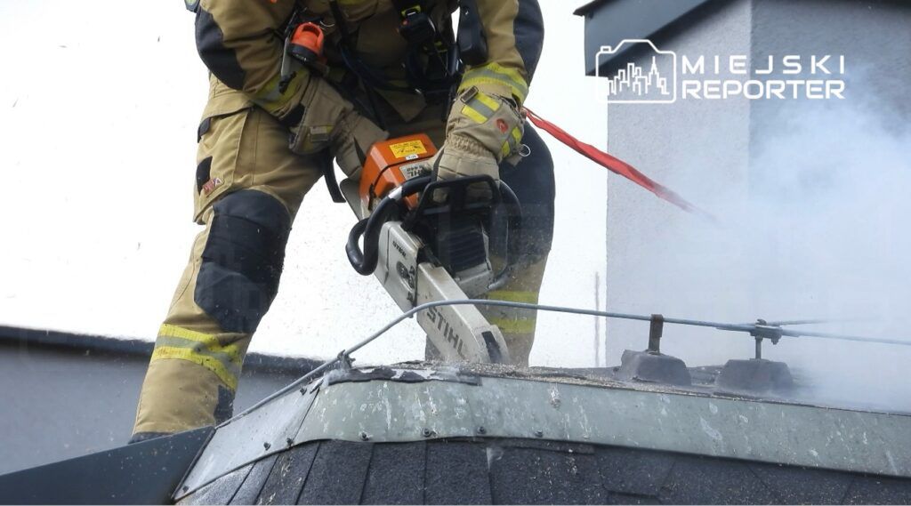 Strażak w mundurze używa piły łańcuchowej do usuwania materiałów z dachu budynku, wokół unosi się dym.