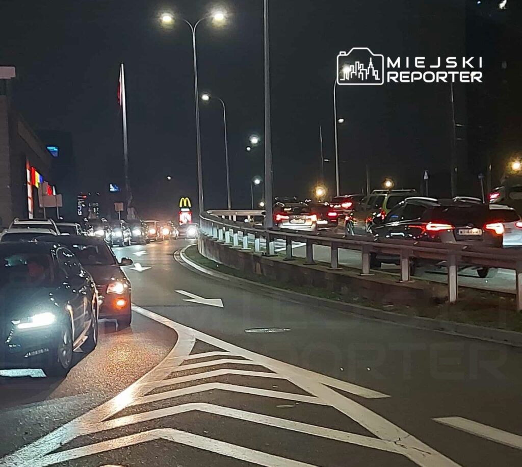 Ruch samochodowy na oświetlonej drodze w nocy, obok restauracji fast food z dużym znakiem.