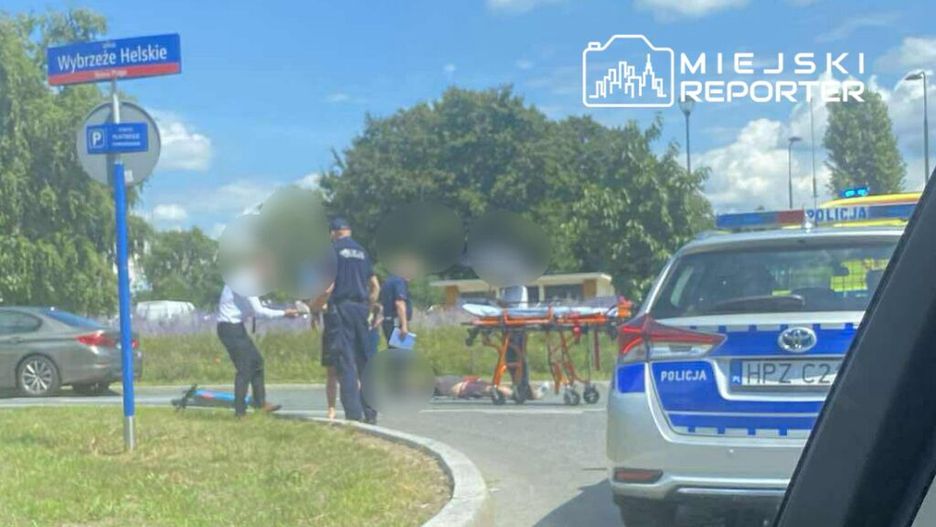 Mężczyzna w garniturze rozmawia z policjantem obok leżącego na ziemi mężczyzny, w tle widać radiowóz.